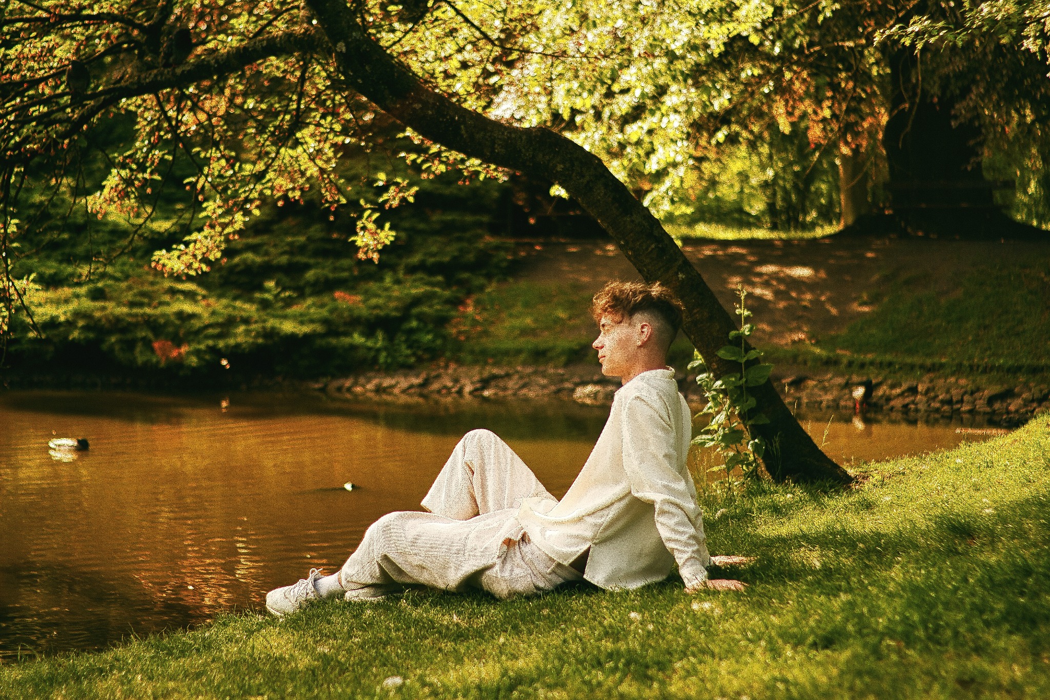 Młody mężczyzna w jasnym ubraniu siedzi na trawie nad brzegiem stawu w parku, z drzewem pochylonym nad wodą, w tle widać zieleń i kaczki pływające po wodzie.