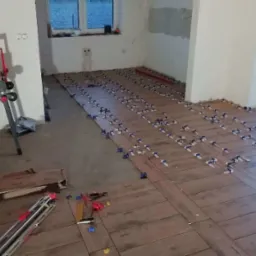 Układanie brązowych płytek podłogowych imitujących drewno z użyciem klipsów poziomujących w trakcie remontu mieszkania, widoczna przecinarka do płytek.