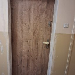 Marcin Tubacki Mar-Art - Zamknięte, drewnopodobne drzwi wewnętrzne z nałożoną gładzią na ościeżnicy i ścianie. Klamka zabezpieczona taśmą malarską. Widoczny fragment ściany z włącznikiem.