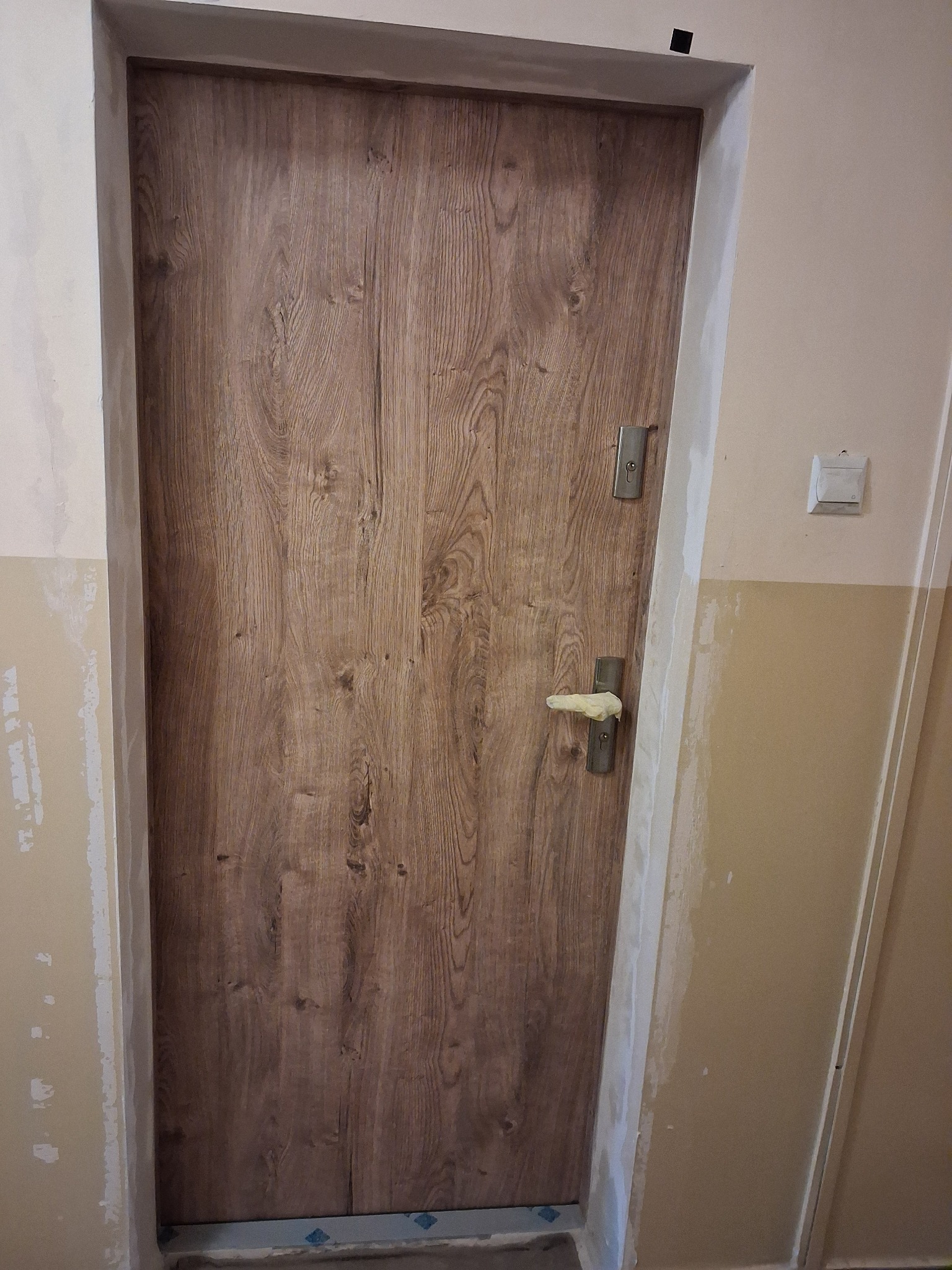 Zamknięte, drewnopodobne drzwi wewnętrzne z nałożoną gładzią na ościeżnicy i ścianie. Klamka zabezpieczona taśmą malarską. Widoczny fragment ściany z włącznikiem.