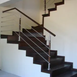 Ciemne drewniane schody z nowoczesną balustradą ze stali nierdzewnej, widok z dołu, minimalistyczny wystrój, kremowe ściany, doniczka w rogu.