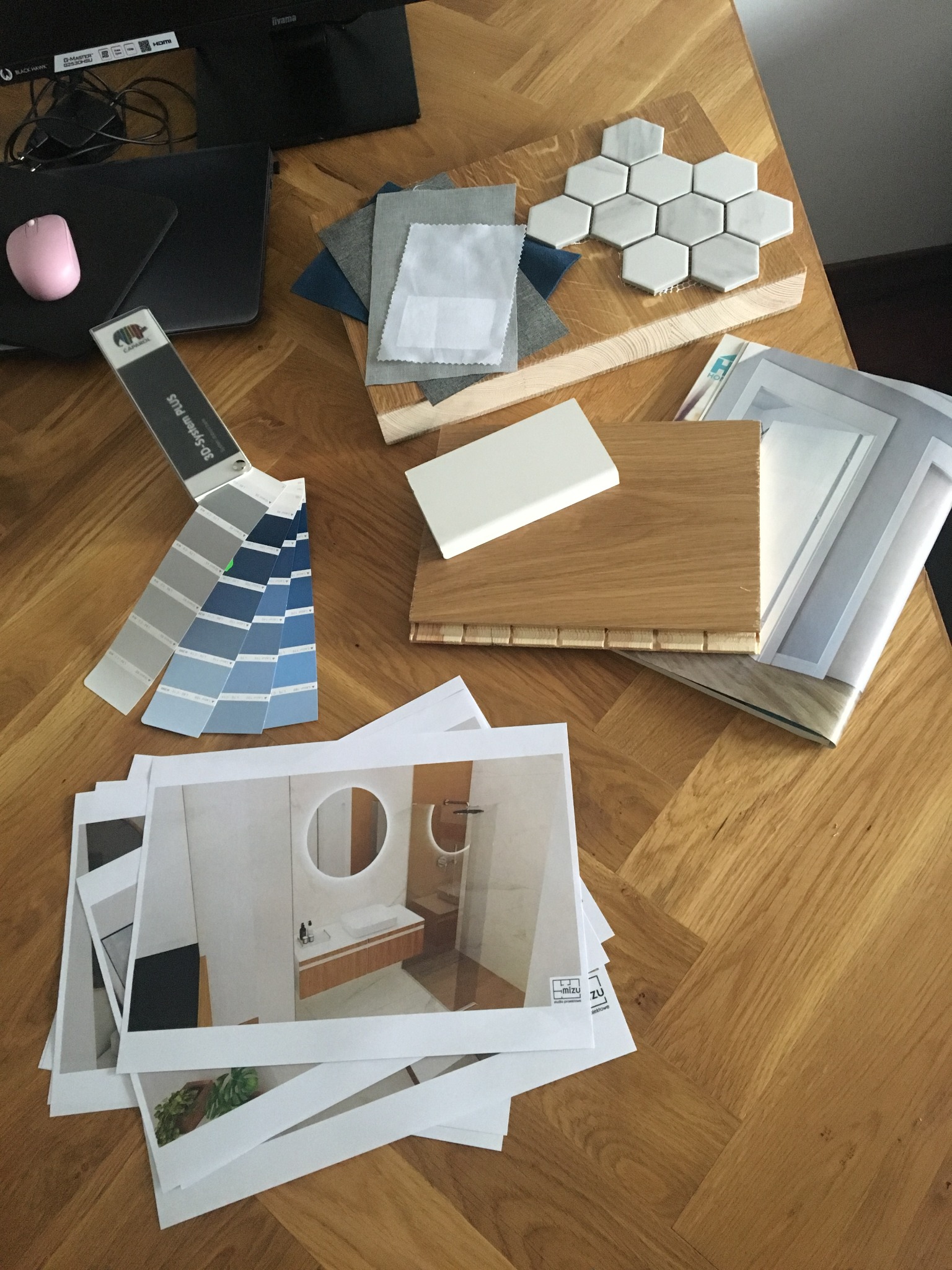 Paleta kolorów, próbki tkanin, płytek heksagonalnych i wizualizacje łazienki ułożone na parkiecie w jodełkę francuską, obok laptop i różowa myszka.