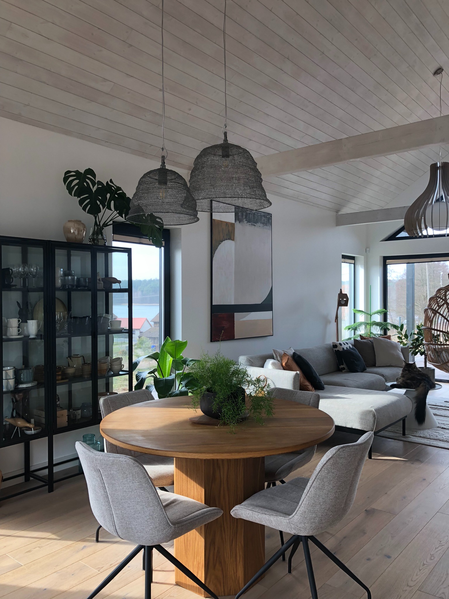 Jasne wnętrze domu z drewnianą podłogą, okrągłym stołem z rośliną, szarymi krzesłami, przeszkloną szafką z dekoracjami, szarą sofą i dużym abstrakcyjnym obrazem na ścianie.