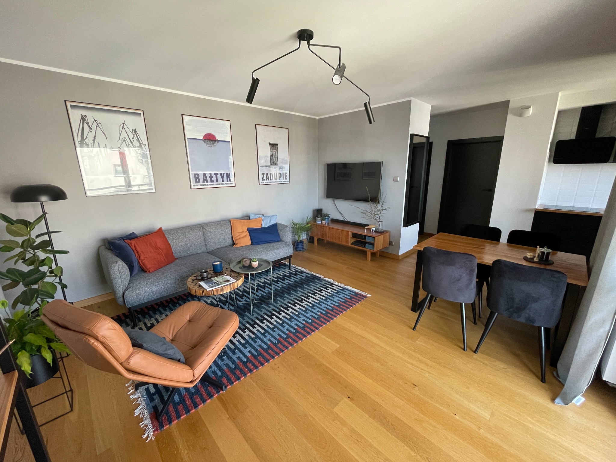 Jasne wnętrze mieszkania z drewnianą podłogą, szarą sofą z kolorowymi poduszkami, brązowym fotelem, dywanem w geometryczne wzory i trzema plakatami na ścianie.