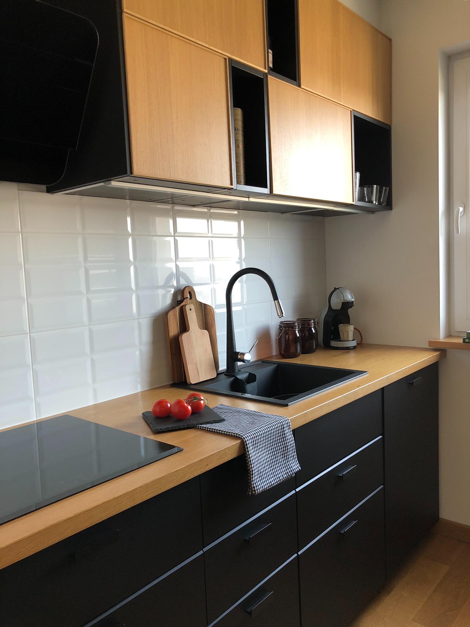 Nowoczesna kuchnia z czarnymi matowymi szafkami dolnymi i jasnymi drewnianymi szafkami górnymi, czarnym zlewem i armaturą, drewnianym blatem, białymi płytkami na ścianie oraz dodatkami: drewniane...