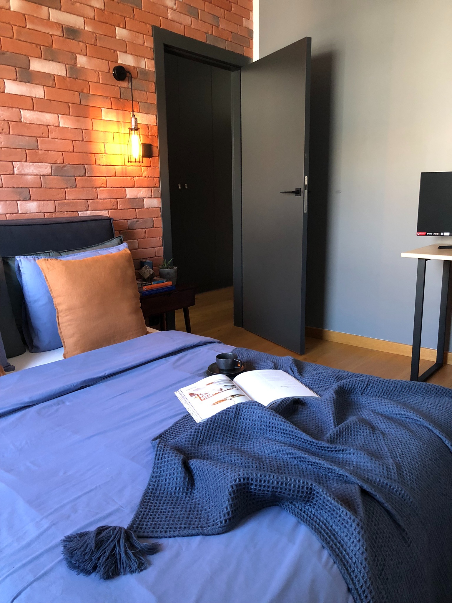 Sypialnia z ceglaną ścianą, industrialną lampą, łóżkiem z niebieską pościelą i książką oraz filiżanką na wierzchu, z czarnymi drzwiami w tle.