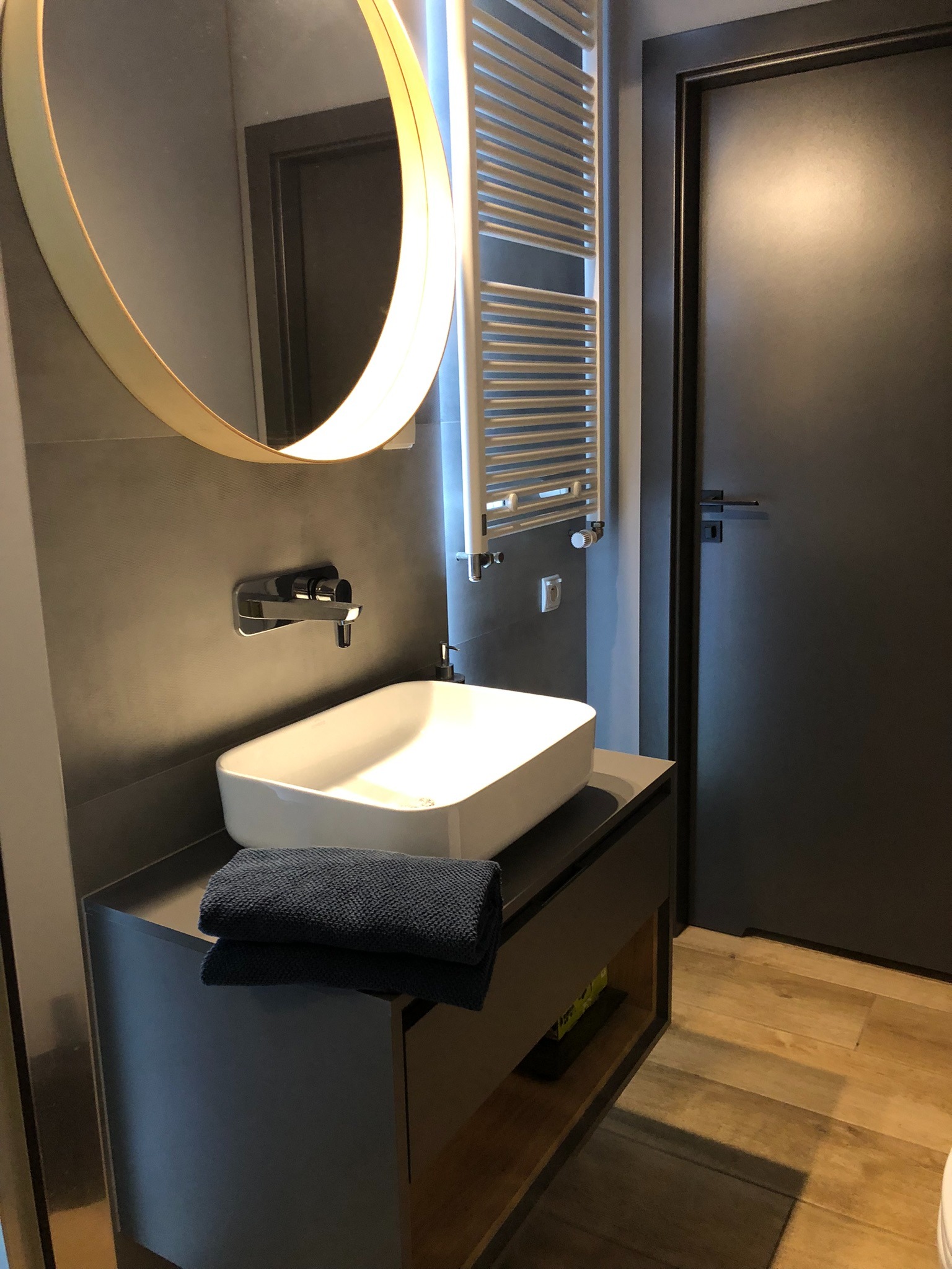 Nowoczesna łazienka w odcieniach szarości z okrągłym lustrem z podświetleniem LED, białą umywalką nablatową i grzejnikiem drabinkowym.