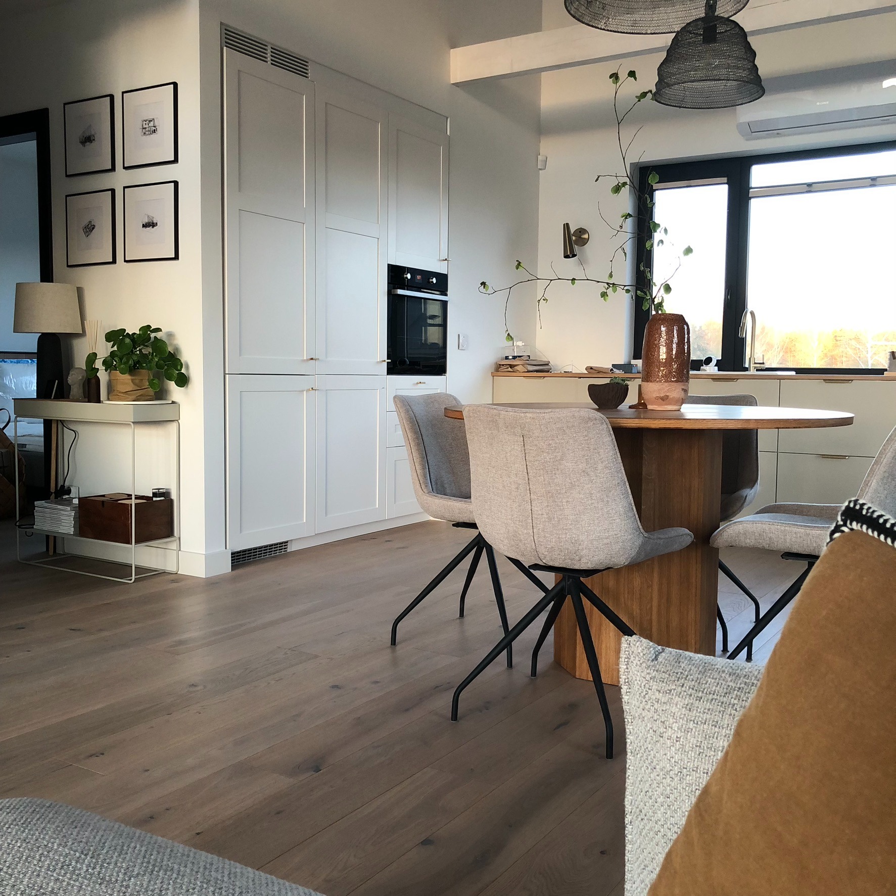 Jasne wnętrze jadalni z okrągłym drewnianym stołem, szarymi krzesłami na czarnych nogach, białymi szafkami kuchennymi i drewnianą podłogą; minimalistyczny wystrój z dekoracjami roślinnymi i lampami...