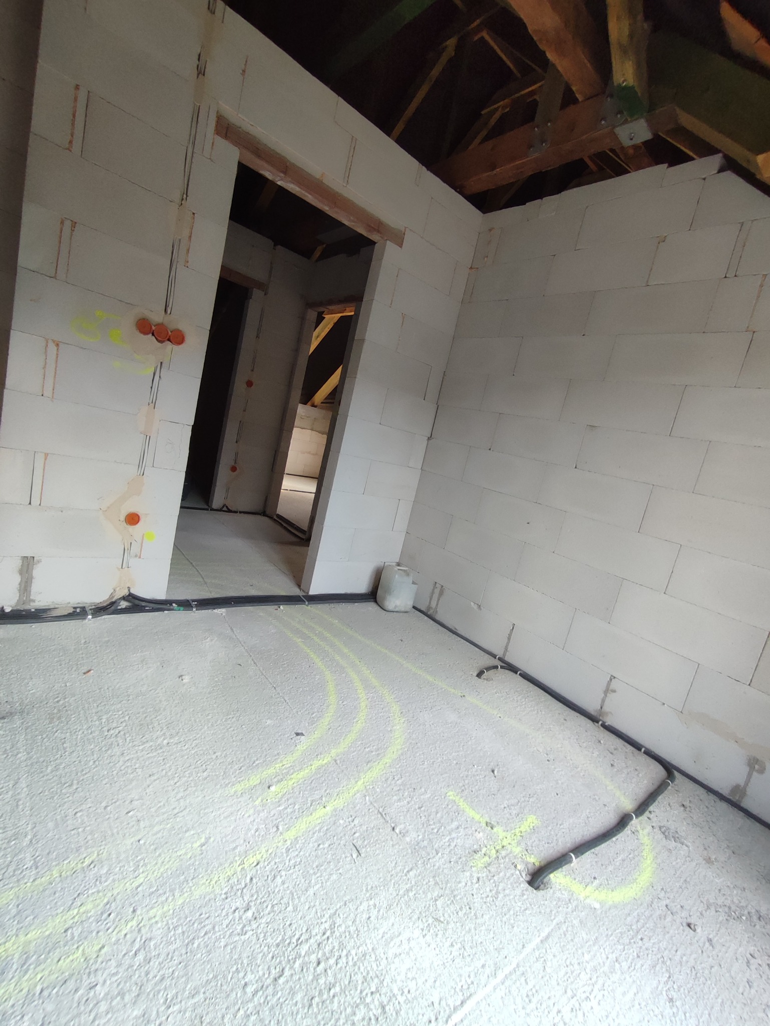 Surowy stan poddasza z instalacją elektryczną w peszlach, widoczne oznaczenia tras kabli żółtym markerem na posadzce, ściany z białych bloczków, widoczne puszki instalacyjne.