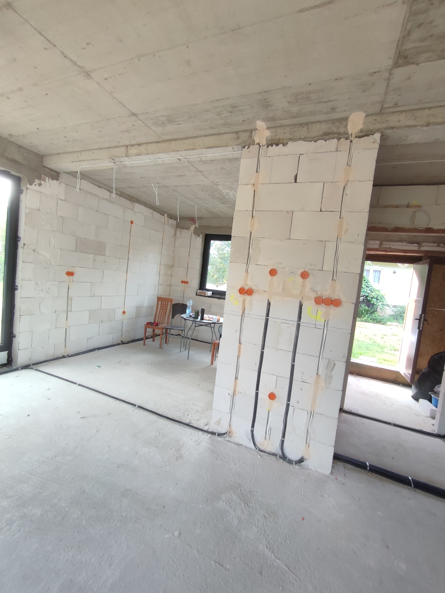 Wnętrze w stanie surowym z widocznymi przewodami elektrycznymi w ścianach z betonu komórkowego, pomarańczowe puszki instalacyjne, czarne kable prowadzone po podłodze, okno i drzwi balkonowe...