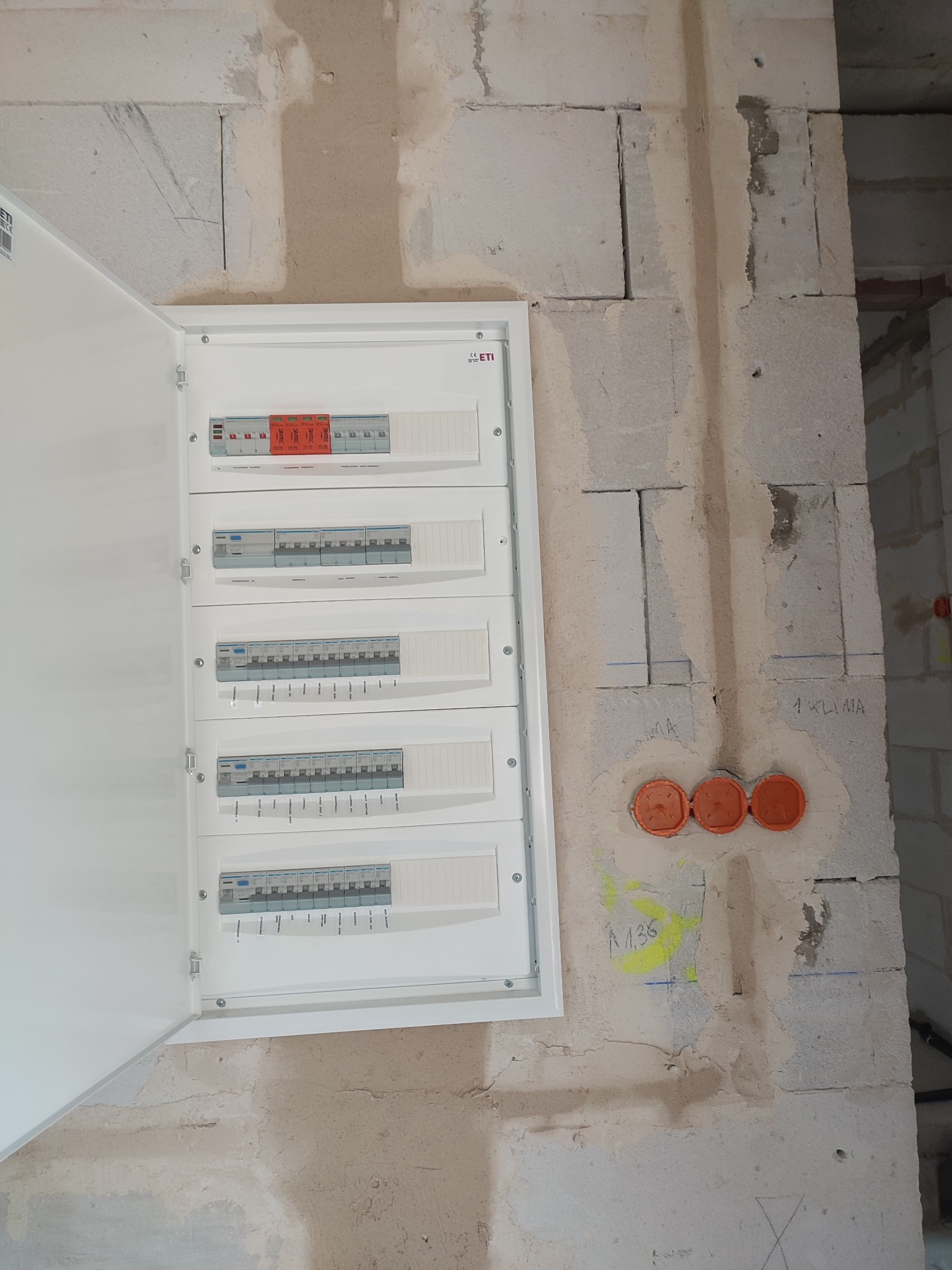 Otwarta skrzynka rozdzielcza ETI z zamontowanymi bezpiecznikami na surowej ścianie z betonu komórkowego, widoczne pomarańczowe puszki instalacyjne i oznaczenia na ścianie.