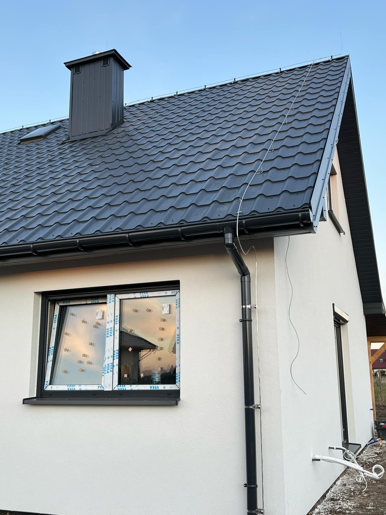 Nowoczesny dom jednorodzinny z grafitową blachodachówką, czarną rynną, białymi ścianami i widoczną instalacją odgromową z przewodem biegnącym wzdłuż dachu i ściany. Okna z folią ochronną.