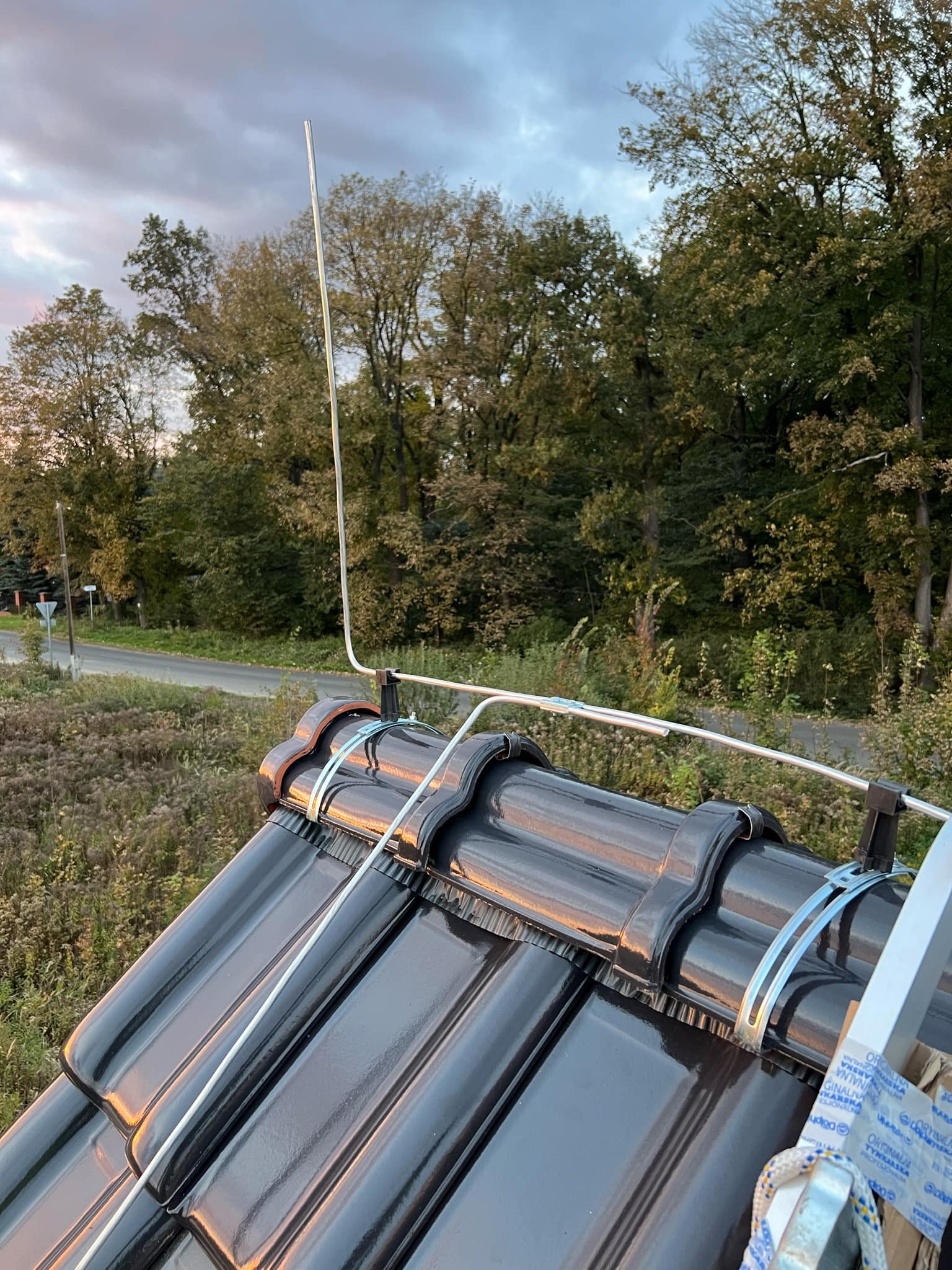 Zmontowana instalacja odgromowa na dachu z czarnej dachówki w Nawojowej, widok z góry na system przewodów i maszt uziemiający na tle drzew i drogi.