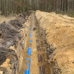 Długi wykop w ziemi w lesie, z widocznymi niebieskimi rurami ułożonymi na dnie i zabezpieczonymi ścianami z desek.