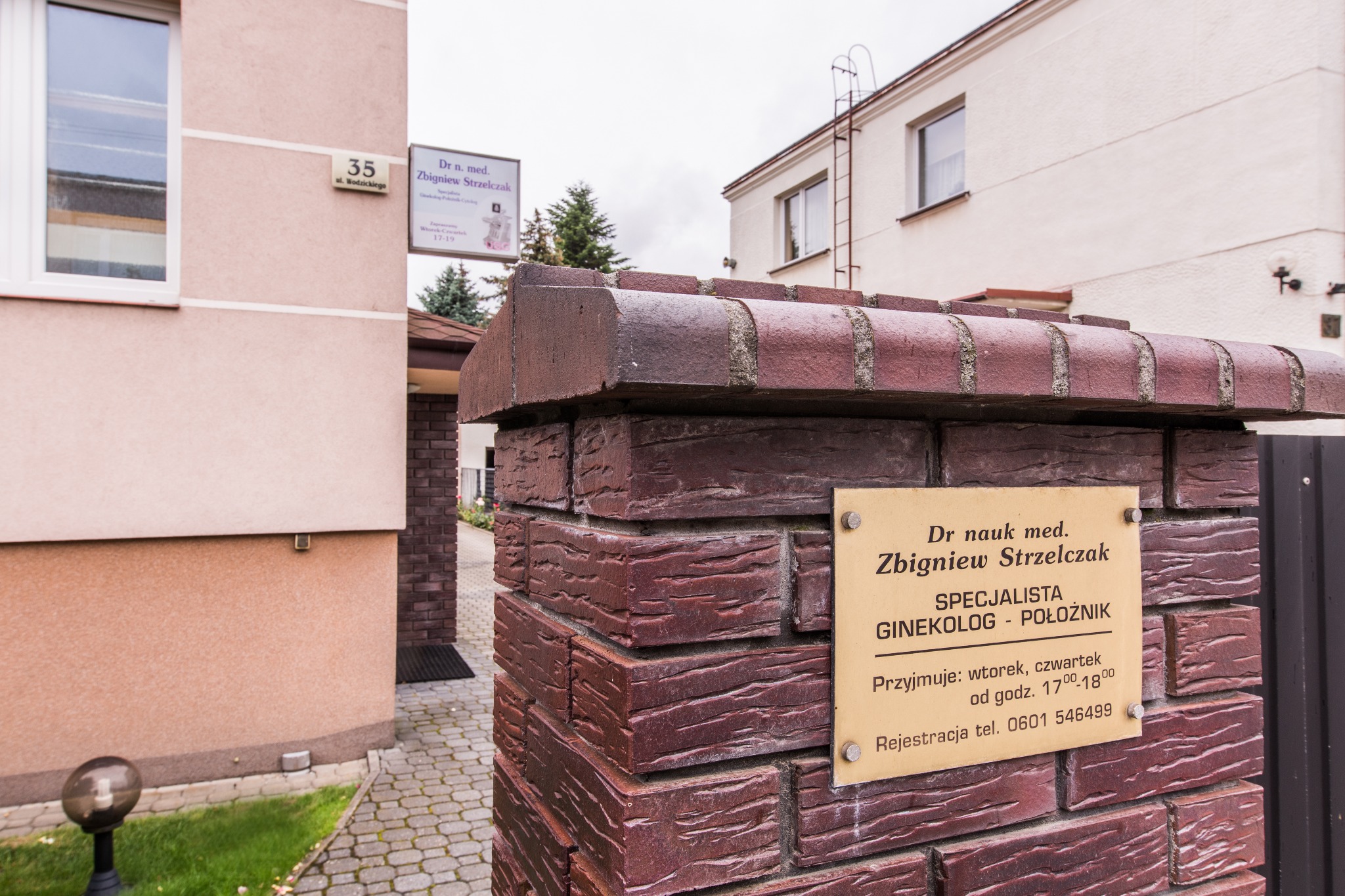 Tabliczka informacyjna z nazwiskiem lekarza specjalisty ginekologa-położnika Zbigniewa Strzelczaka umieszczona na ceglanym murku przed budynkiem w Częstochowie. Widoczny adres i godziny przyjęć.