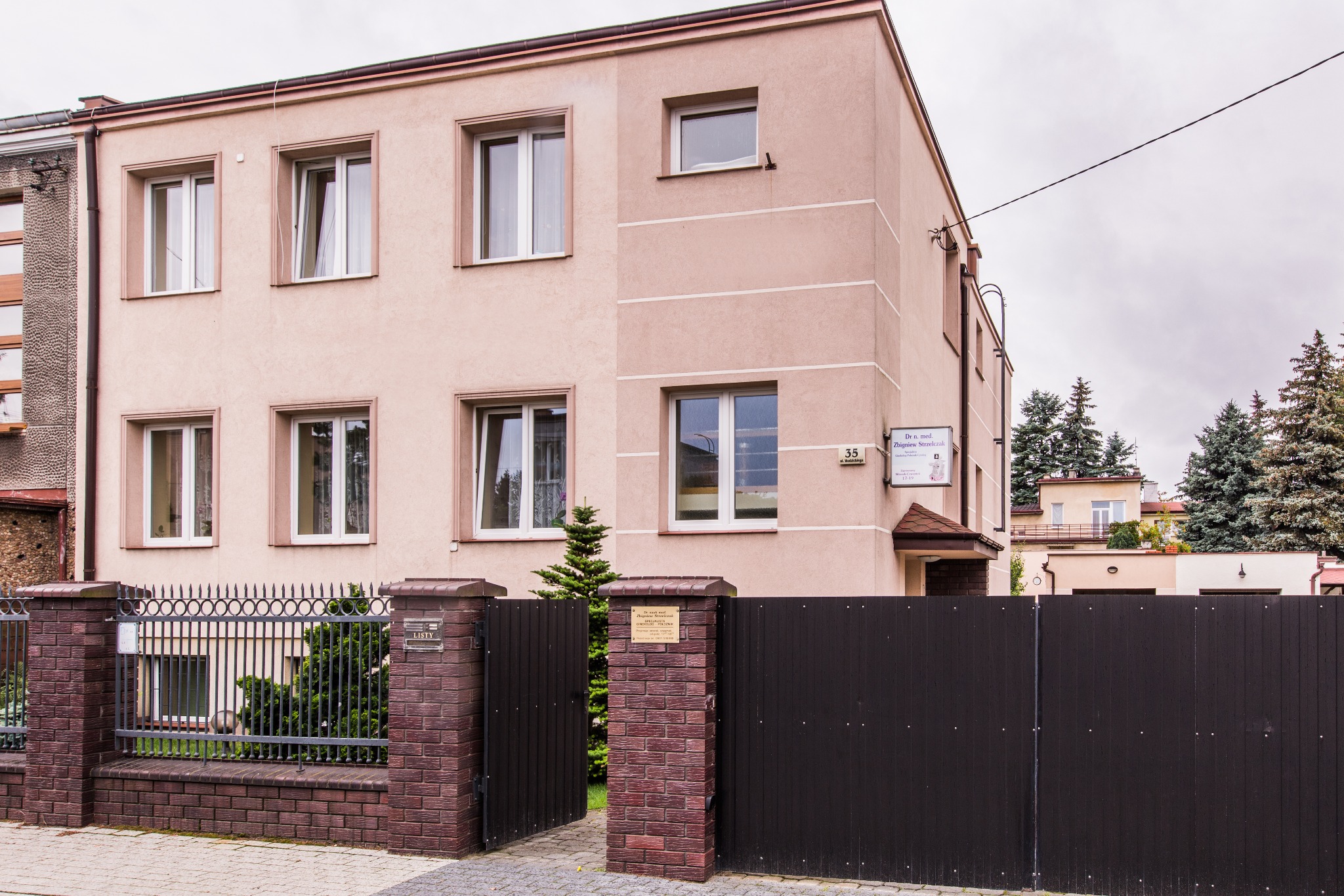 Budynek gabinetu lekarskiego z tabliczką informacyjną na elewacji, widoczny zza ogrodzenia z bramą