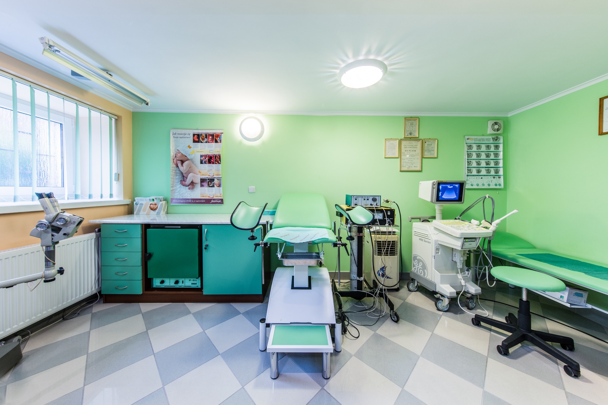 Jasne wnętrze gabinetu lekarskiego z zielonymi ścianami, wyposażone w fotel ginekologiczny z podnóżkiem, aparat USG na kółkach, mikroskop diagnostyczny i szafki medyczne.