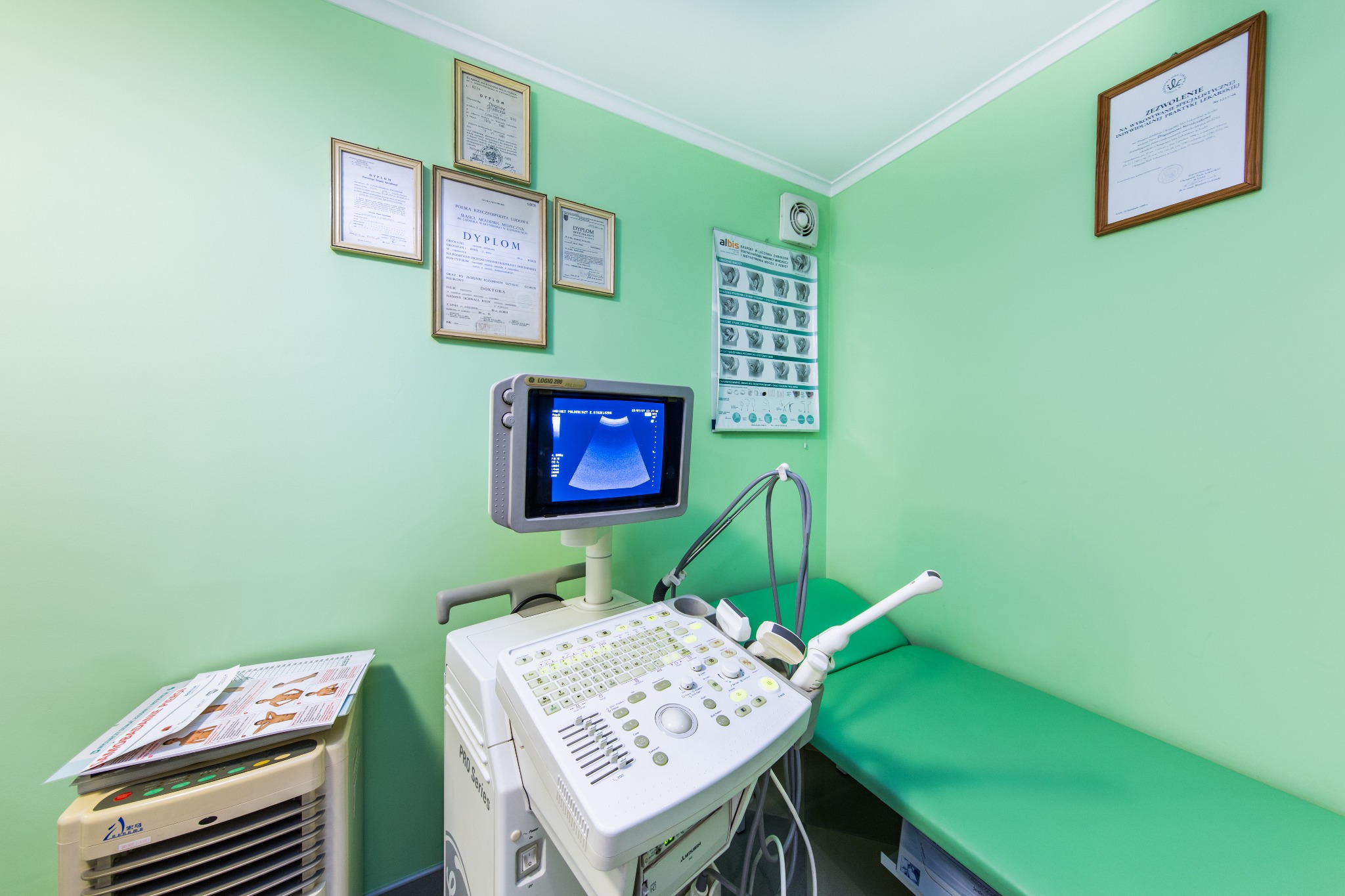 Gabinet ginekologiczny z aparatem USG LOGIQ 200, leżanką i dyplomami na ścianie w odcieniach zieleni.