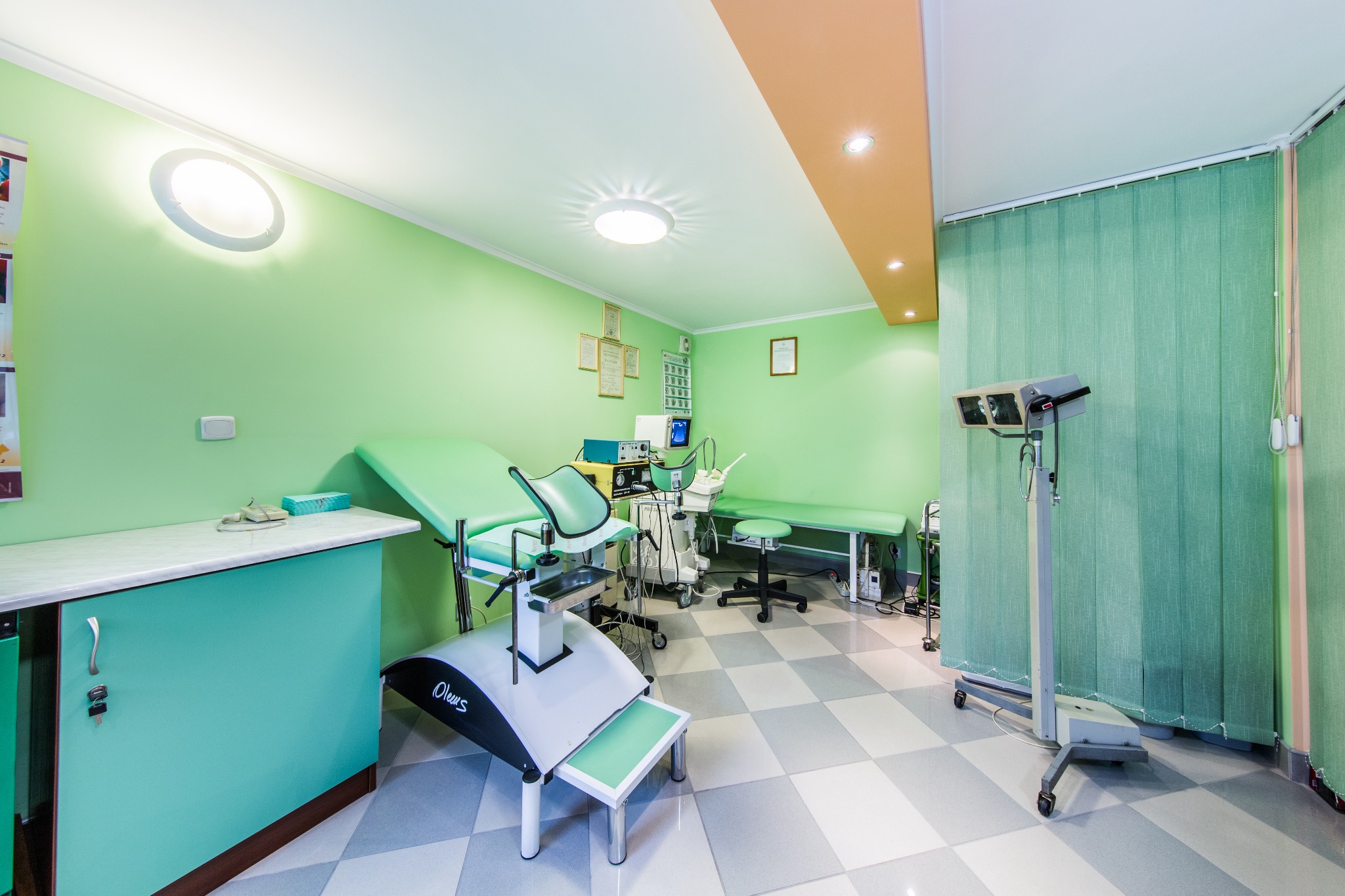 Jasne wnętrze gabinetu lekarskiego z zielonymi ścianami, wyposażone w fotel ginekologiczny, kozetkę, lampę diagnostyczną i sprzęt medyczny na kółkach.