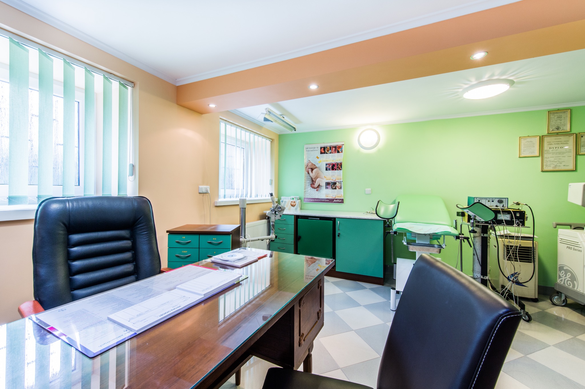 Jasny gabinet lekarski z biurkiem, czarnym fotelem, zielonymi szafkami i sprzętem medycznym; na ścianie dyplomy i plakat informacyjny.