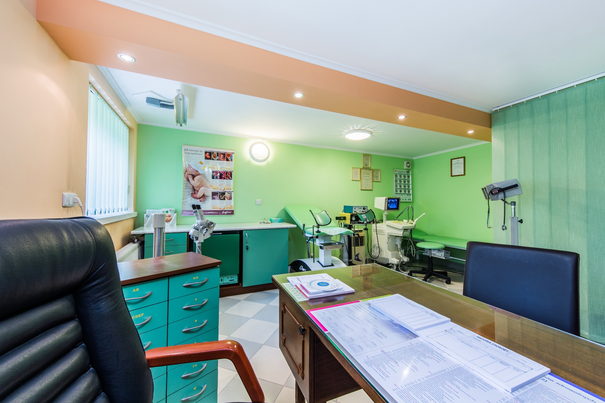Gabinet lekarski z zielonymi ścianami, biurkiem z dokumentami medycznymi i aparaturą diagnostyczną w tle.