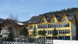 Żółty budynek z ciemnym dachem i trzema szczytami, otoczony śniegiem i drzewami iglastymi w górskim krajobrazie pod błękitnym niebem.