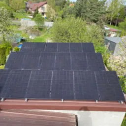 Widok z góry na zainstalowane panele fotowoltaiczne na dachu domu jednorodzinnego, z kwitnącymi drzewami i zabudową w tle.