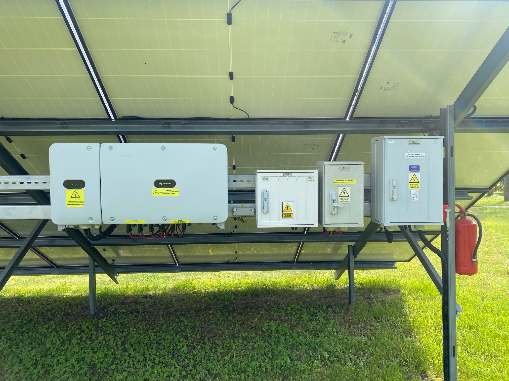 Widok od spodu na panele słoneczne z widocznymi inwerterami Huawei i skrzynkami elektrycznymi, przymocowanymi do metalowej konstrukcji, z trawiastym podłożem w tle i czerwoną gaśnicą po prawej...