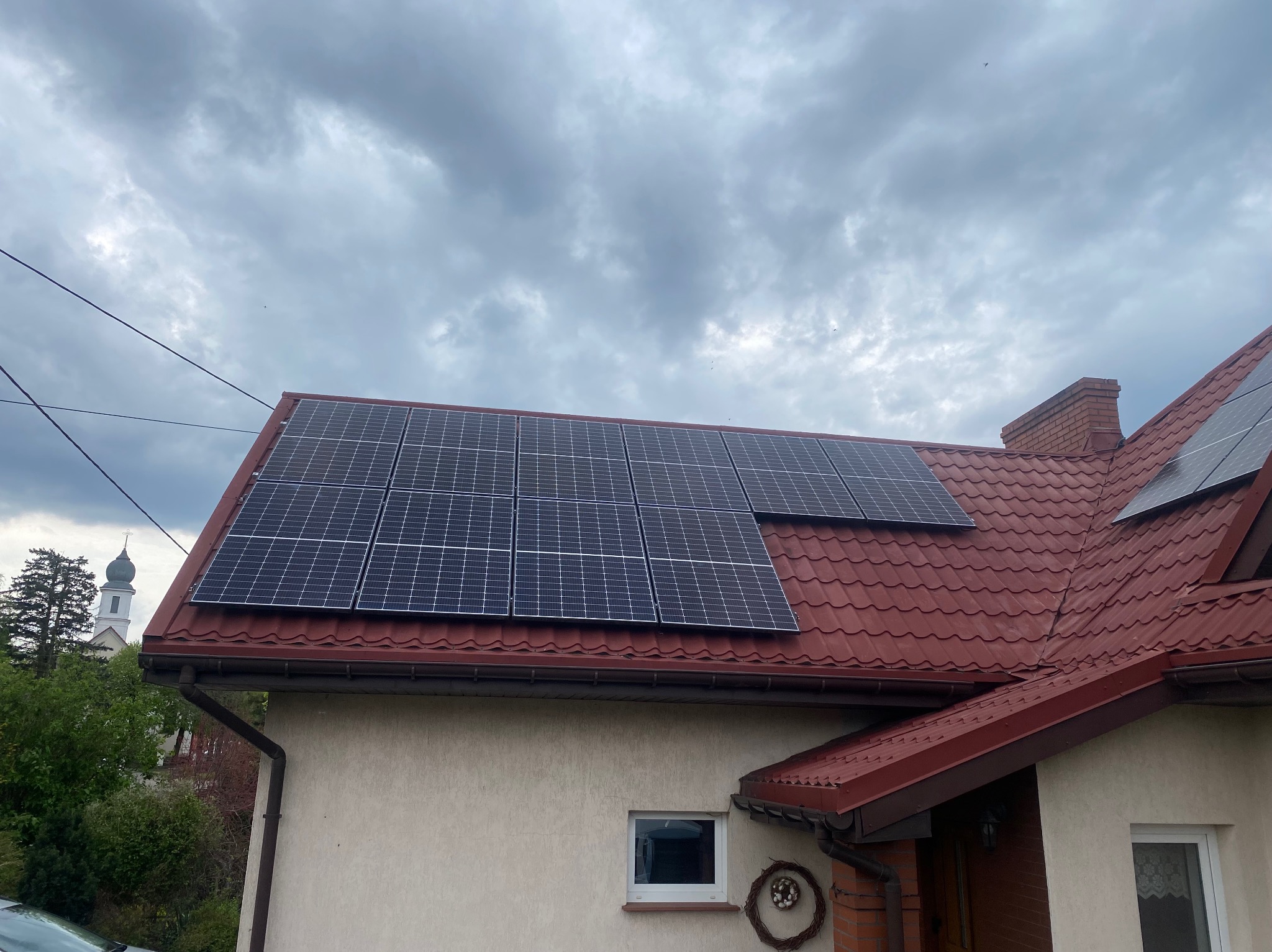 Panele słoneczne na czerwonym dachu domu jednorodzinnego, widok z dołu na pochmurne niebo, w tle wieża kościoła.