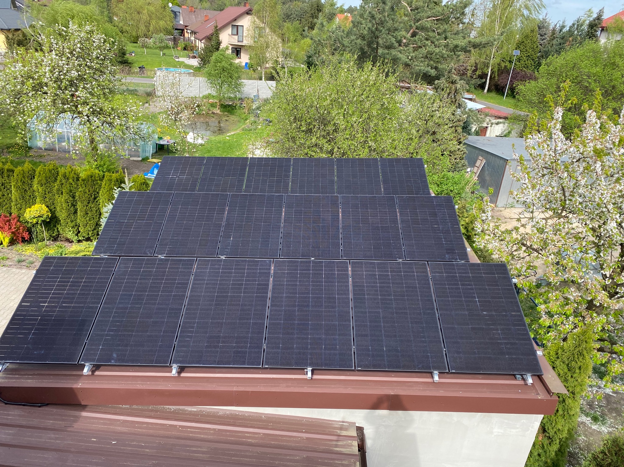 Widok z góry na zainstalowane panele fotowoltaiczne na dachu domu jednorodzinnego, z kwitnącymi drzewami i zabudową w tle.