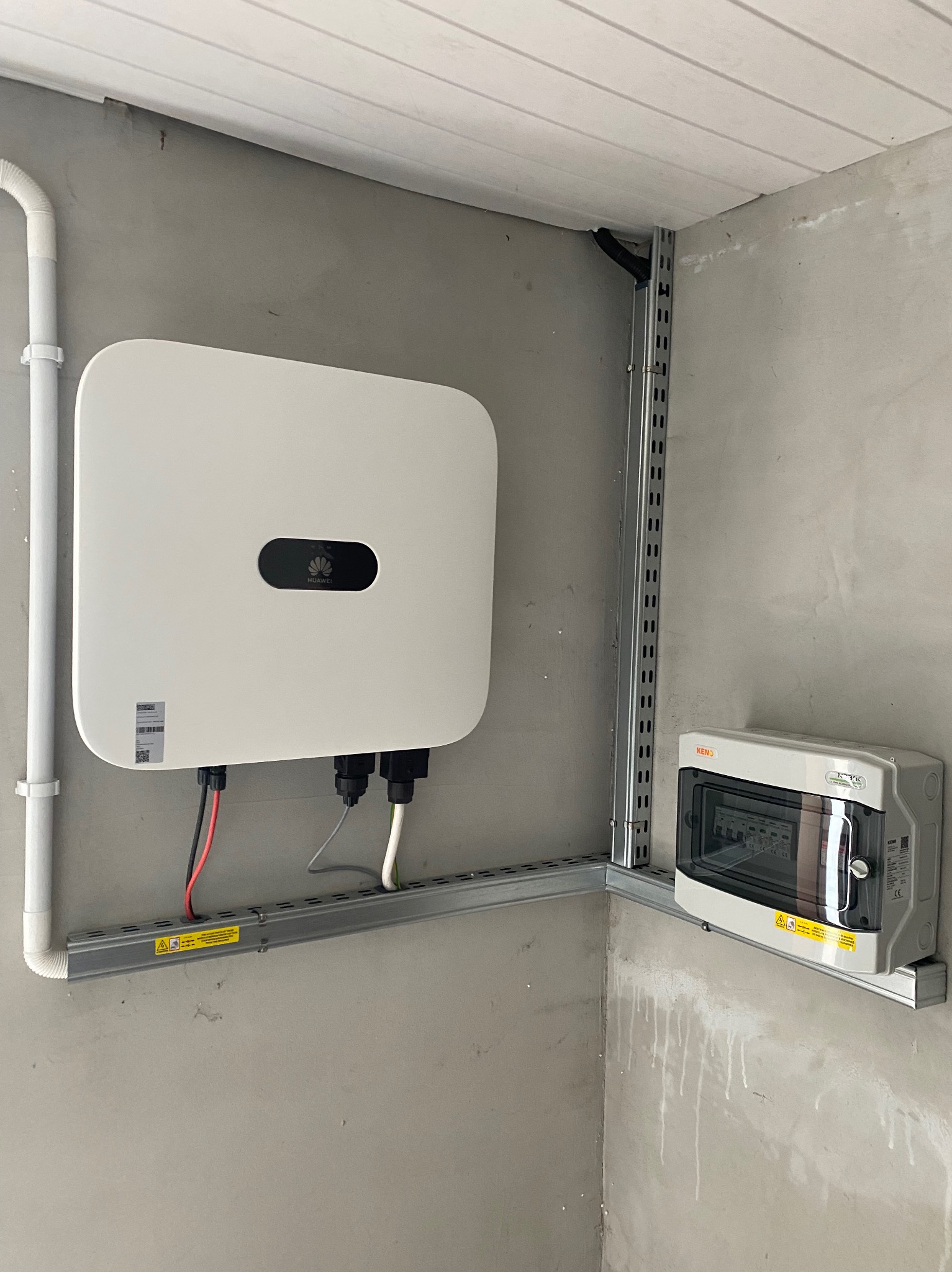 Biały inwerter z logo na szarej ścianie nad listwą montażową, obok szara skrzynka bezpiecznikowa z przezroczystym okienkiem, zamontowane na metalowych profilach.