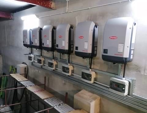 Rząd szarych falowników marki Fronius zamontowanych na szynie w surowym, betonowym pomieszczeniu, z widocznymi przewodami i skrzynkami elektrycznymi, sugerujący instalację systemu.