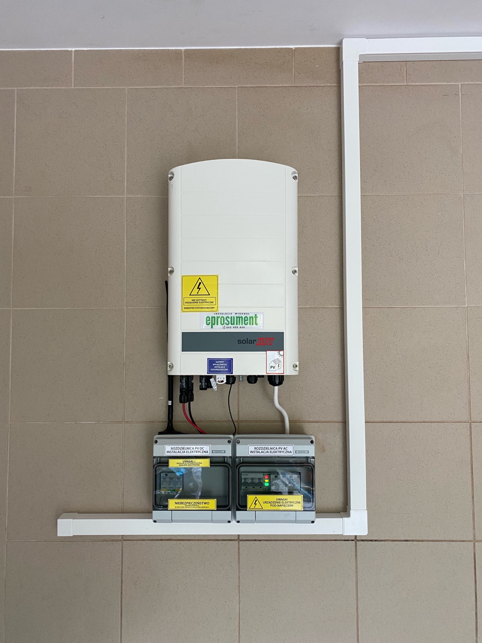 Biały inwerter fotowoltaiczny marki SolarEdge z podłączonymi rozdzielnicami PV DC i PV AC, zamontowany na ścianie wyłożonej beżowymi płytkami, zabezpieczony białymi listwami przypodłogowymi.