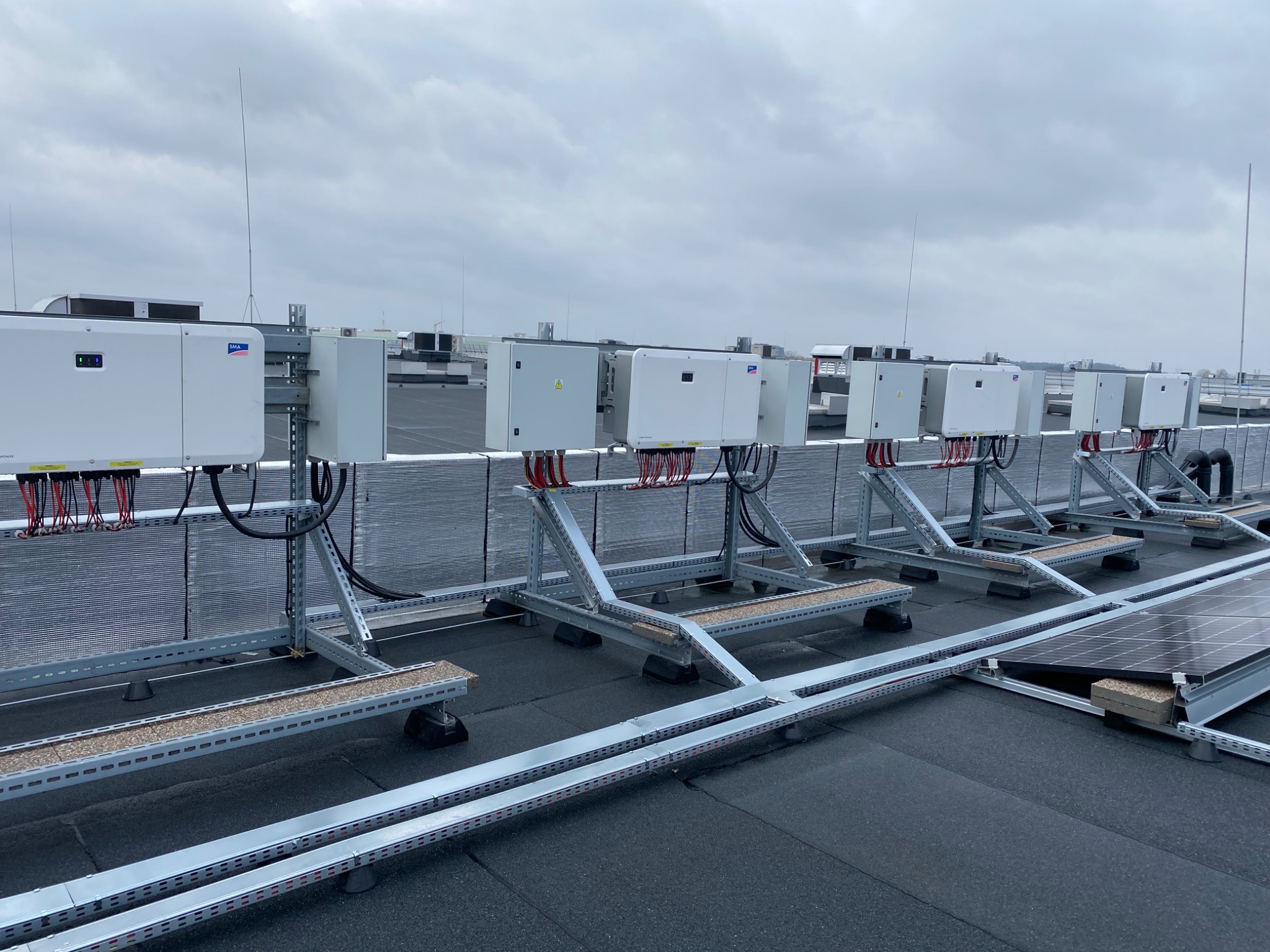 Rząd białych falowników zamontowanych na metalowej konstrukcji na dachu z panelami słonecznymi w tle, widok z góry na pochmurny dzień.