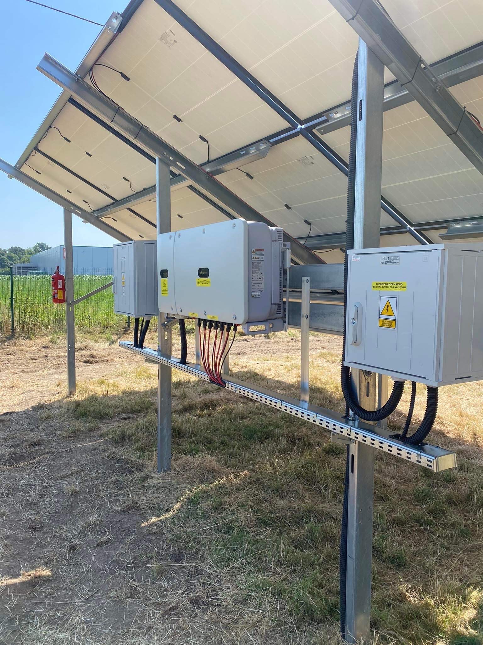 Panele słoneczne zamontowane na metalowej konstrukcji z widocznymi inwerterami i skrzynkami przyłączeniowymi, na tle trawy i budynku magazynowego.