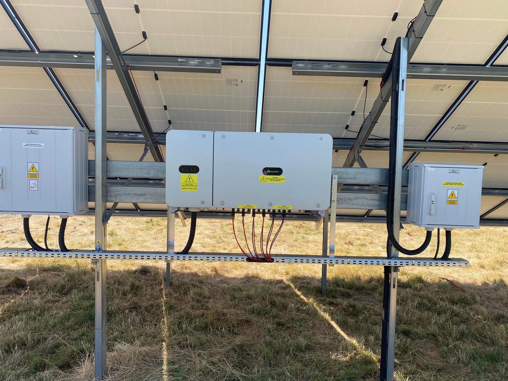 Widok od spodu na panele słoneczne z widocznymi skrzynkami przyłączeniowymi i inwerterem Huawei, zamontowanymi na metalowej konstrukcji, na tle trawy.