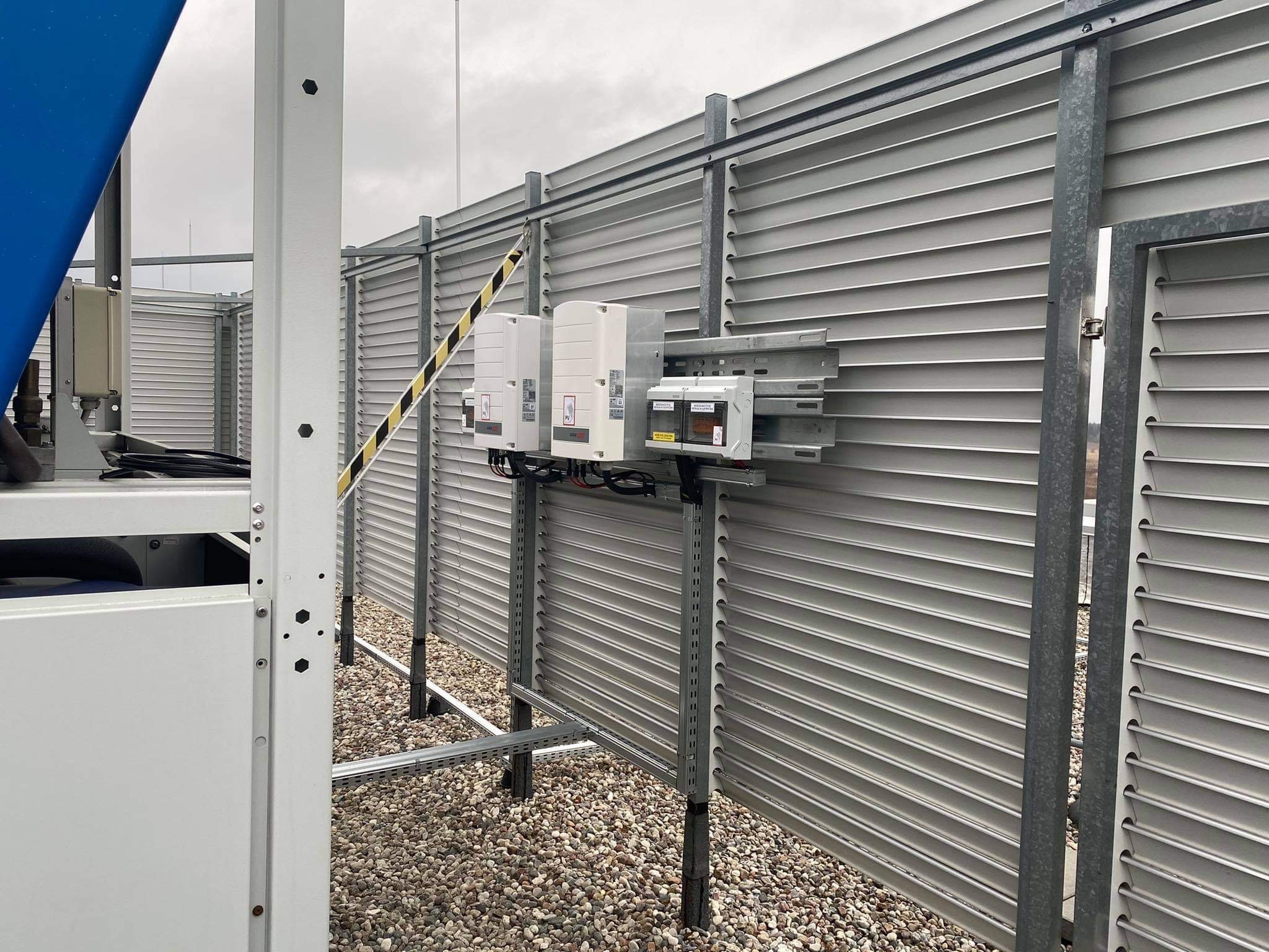 Dwa białe inwertery fotowoltaiczne zamontowane na metalowej konstrukcji na dachu budynku, z widocznymi kablami i elementami instalacji elektrycznej, na tle szarej ściany z żaluzjami.