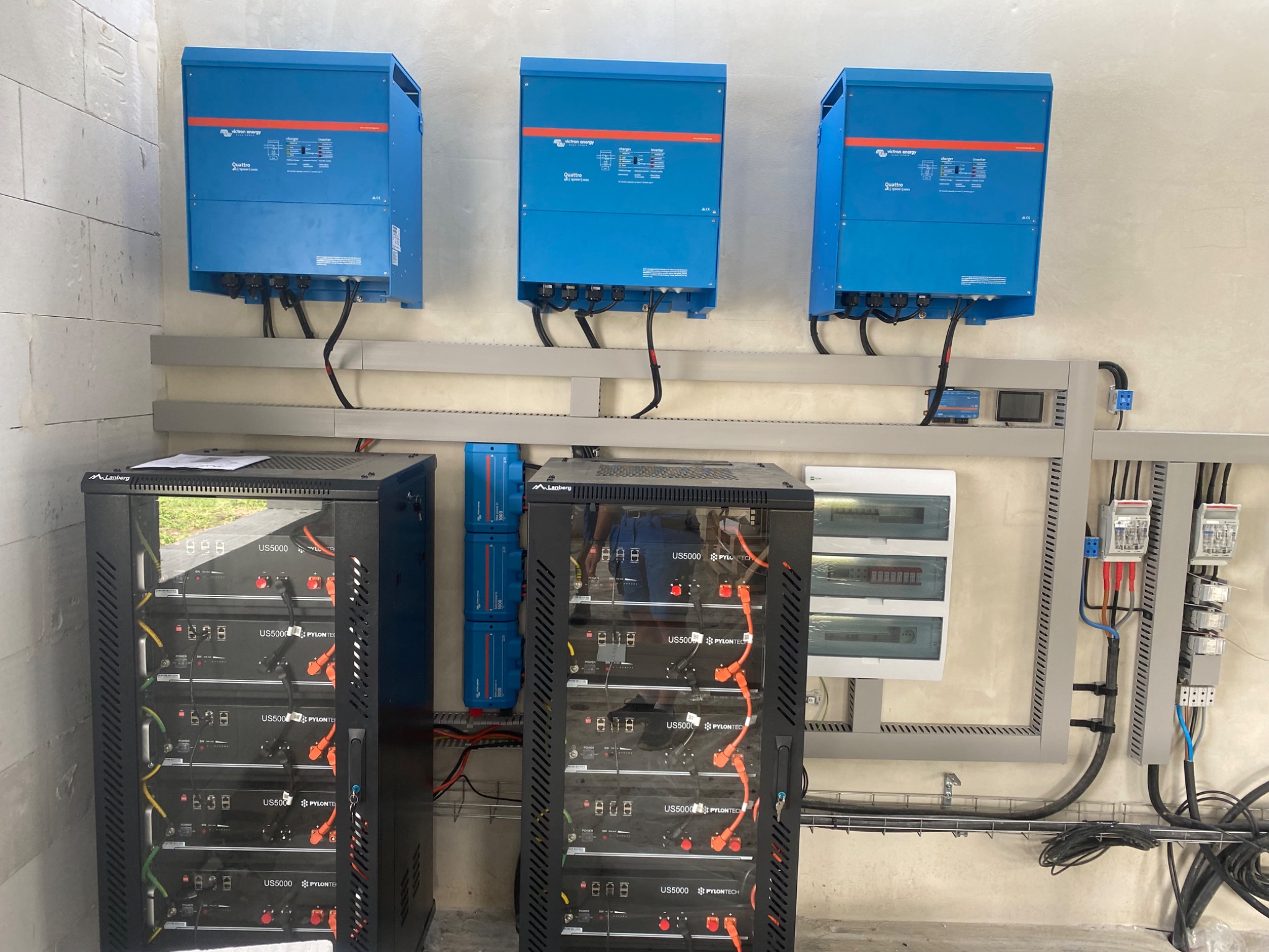 Instalacja z trzema niebieskimi falownikami Victron Energy Quattro i dwoma szafami Lanberg z bateriami Pylontech US5000, okablowanie na szarych listwach, widoczne zabezpieczenia i rozdzielnice.