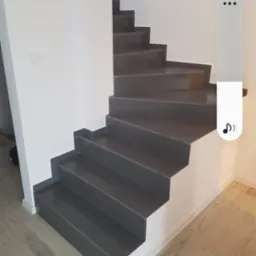 Nowoczesne schody wewnętrzne z ciemnoszarymi stopniami i białymi ścianami, widoczne drewniane panele podłogowe, subtelne oświetlenie.