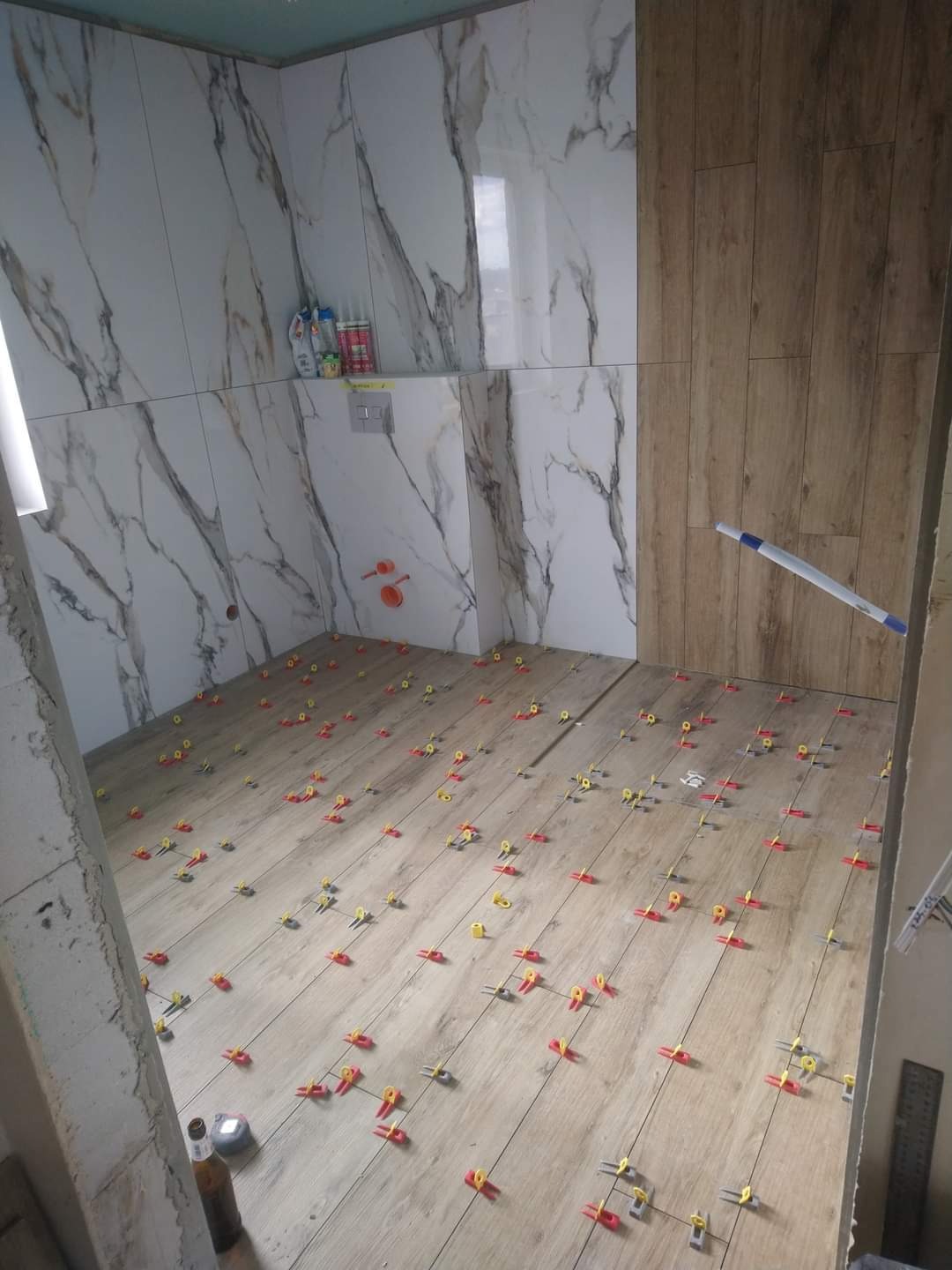 Układanie płytek imitujących drewno na podłodze w łazience, z użyciem czerwono-żółtych klipsów poziomujących, ściany wyłożone płytkami marmurkowymi i drewnopodobnymi.