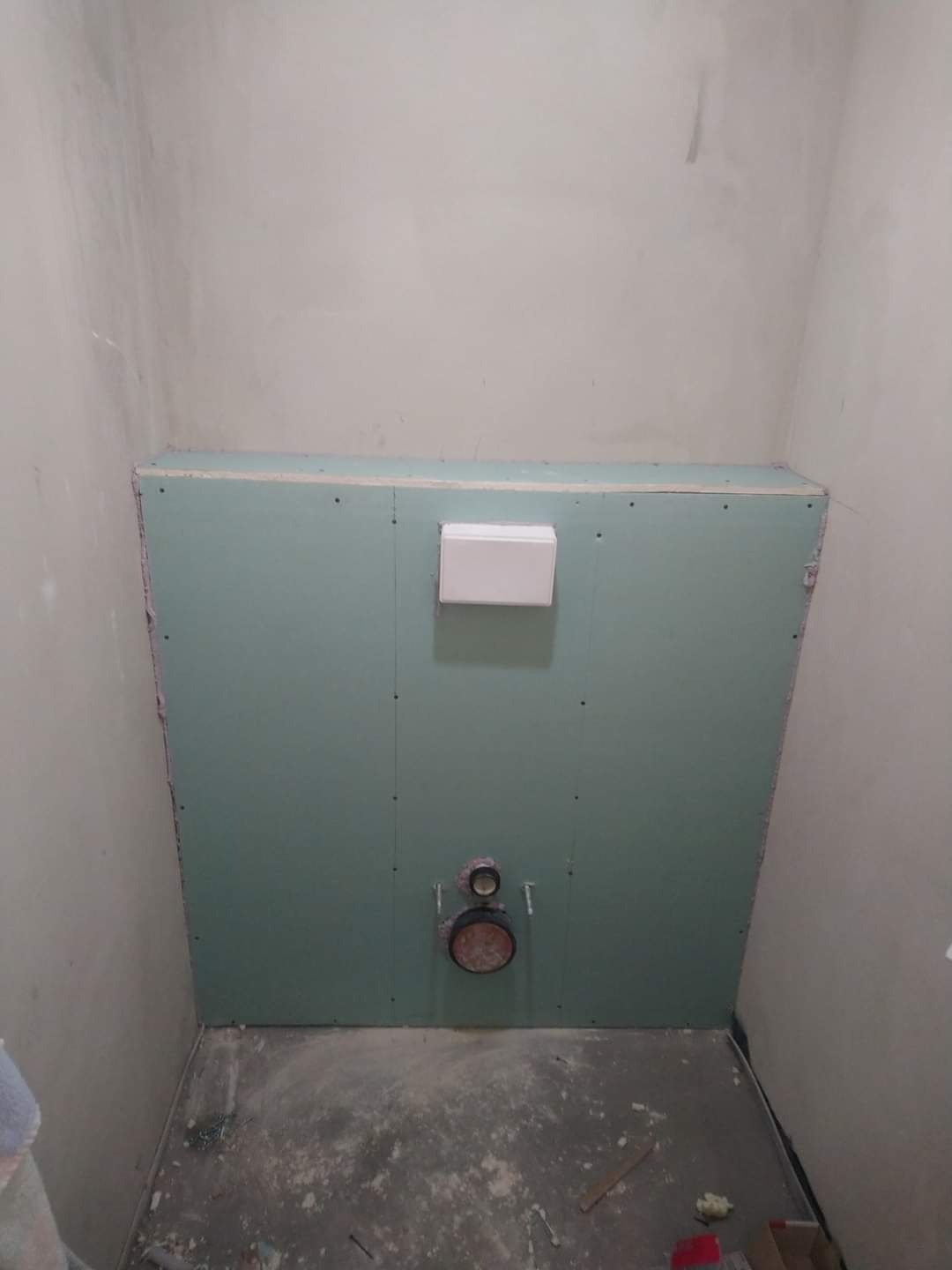 Przygotowanie do montażu stelaża podtynkowego w łazience: płyty gipsowo-kartonowe zamontowane na ścianie, widoczne przyłącza kanalizacyjne i wentylacyjne, surowy beton na podłodze z resztkami...