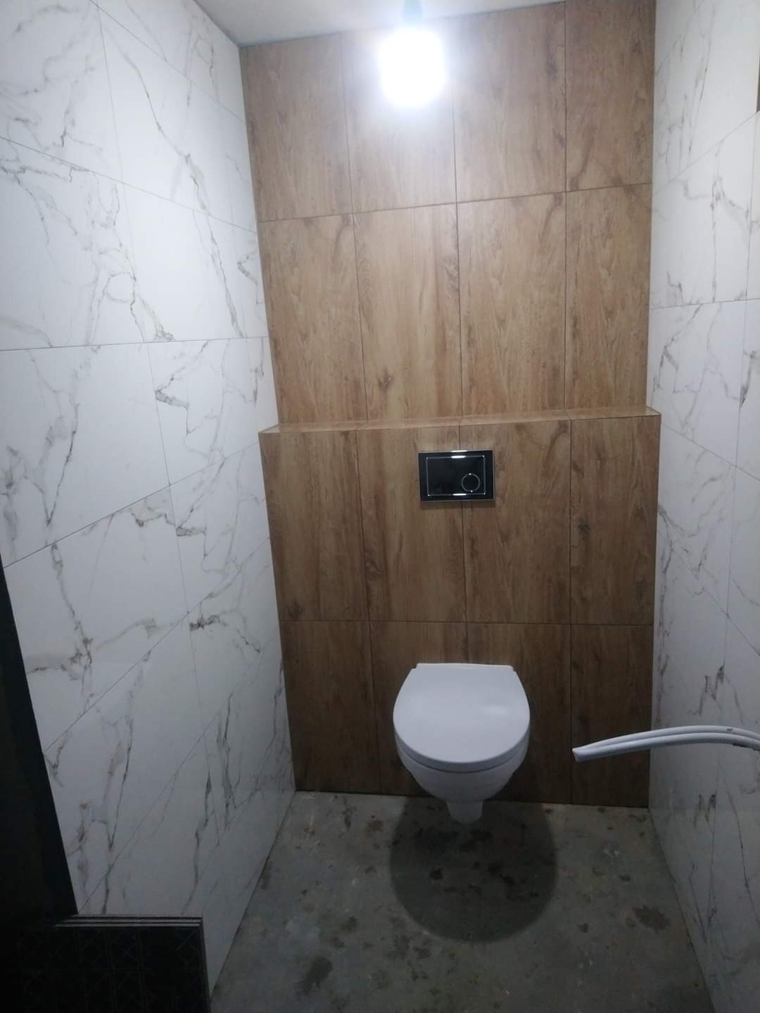 Nowoczesna toaleta z białymi marmurkowymi płytkami na ścianach i drewnopodobnymi płytkami za podwieszaną muszlą klozetową, surowy beton na podłodze, widoczny fragment wężyka.