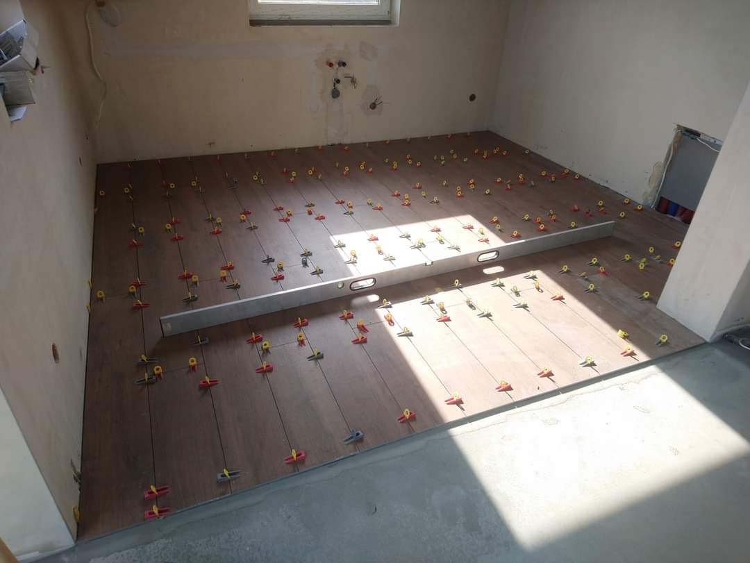 Układanie brązowych płytek podłogowych z użyciem systemu poziomowania, widoczna poziomica i sznurki wyznaczające linie, surowy stan ścian.