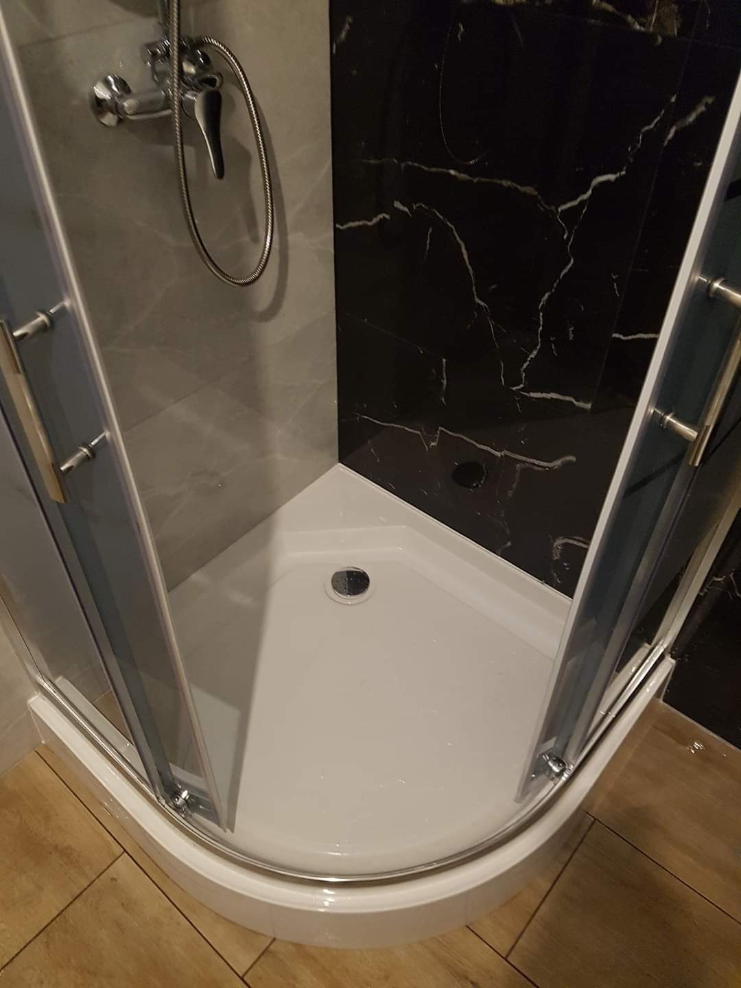 Kabina prysznicowa narożna z brodzikiem, chromowaną armaturą i czarną ścianą z marmurowym wzorem, widoczna z perspektywy podłogi wyłożonej drewnopodobnymi panelami.