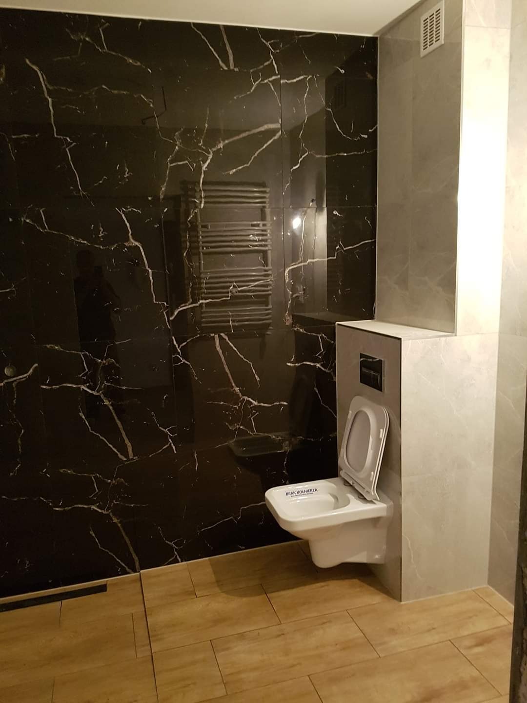 Nowoczesna łazienka z czarnymi płytkami imitującymi marmur, drewnopodobną podłogą i podwieszaną toaletą z otwartą klapą, widoczny grzejnik drabinkowy.