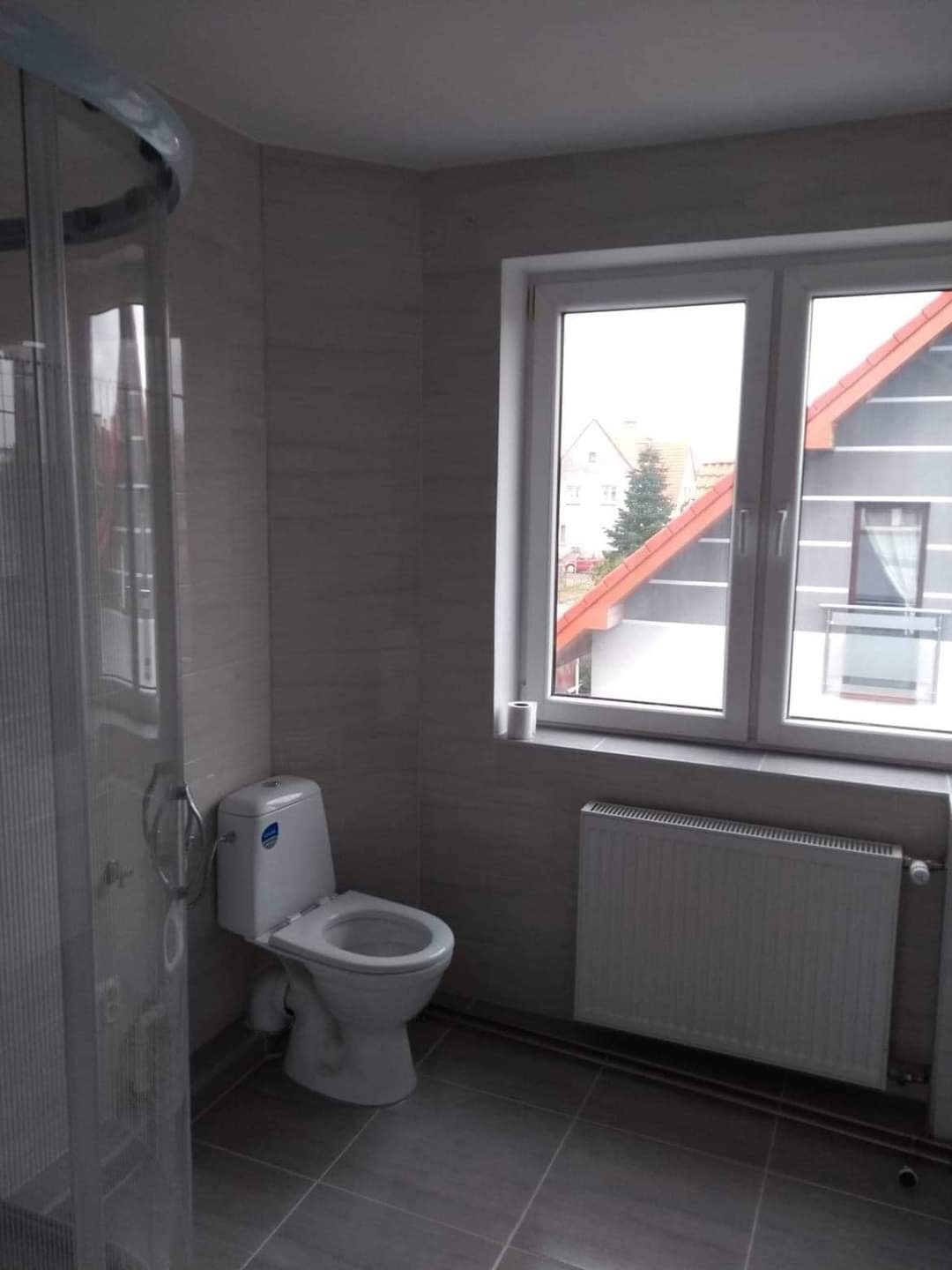 Wykończona łazienka z kabiną prysznicową, toaletą i grzejnikiem pod oknem z widokiem na dom z czerwoną dachówką.