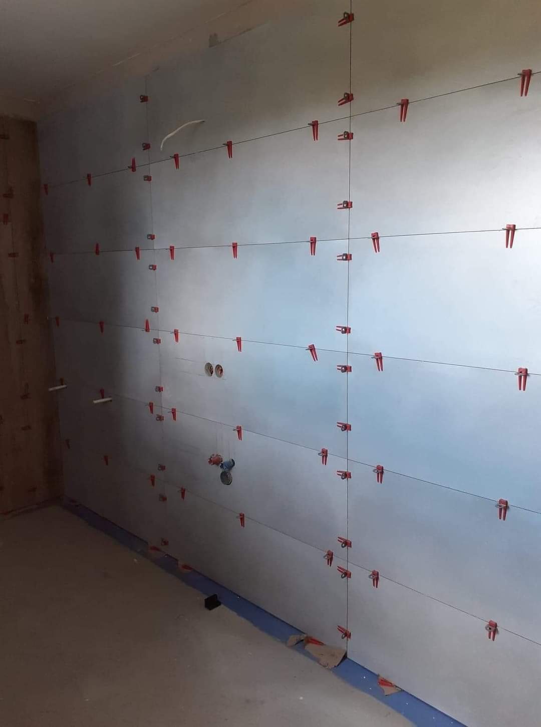 Świeżo ułożone, duże, szare płytki ścienne w łazience, z użyciem czerwonych klipsów poziomujących i czarnej żyłki, widoczne otwory na armaturę.