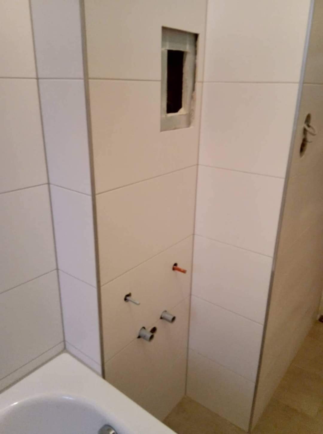 Ściana łazienki w trakcie wykańczania, widoczne białe płytki, otwór na instalację i wystające rurki pod baterię.