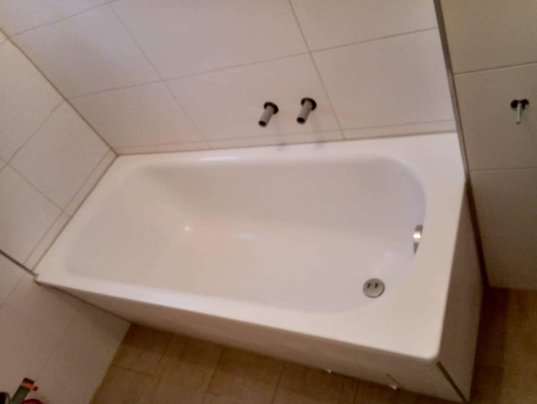 Biała, prostokątna wanna w obudowie, z widocznymi chromowanymi elementami armatury, zamontowana w narożniku wykafelkowanej łazienki z widocznymi przyłączami do baterii.