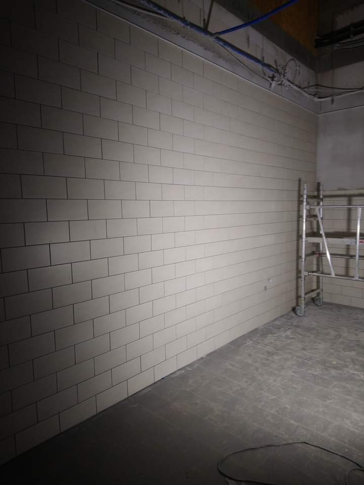 Ściana wyłożona jasnymi, prostokątnymi płytkami ceramicznymi z ciemną fugą, widoczna drabina aluminiowa i fragment surowego sufitu z instalacjami.